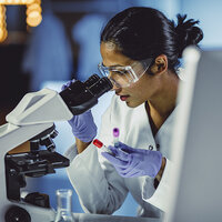 A researcher holding two vials while looking into a microscope. A researcher holding two vials while looking into a microscope.