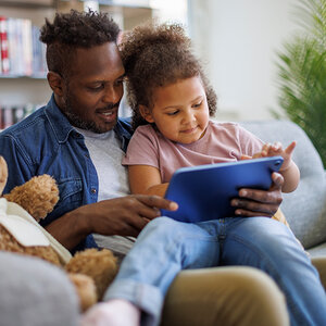 A young girl sits on her father's lap as he reads to her from a book. A young girl sits on her father's lap as he reads to her from a book.