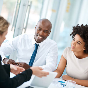 A man and a woman of color talking across a desk to someone in profile. A man and a woman of color talking across a desk to someone in profile.