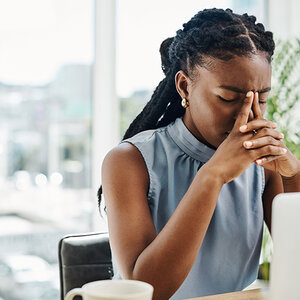 Stressed Black in an office woman at laptop clasps her nose between her fingers.