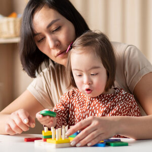 A girl with Down Syndrome playing with the help of a teacher stting behind her.