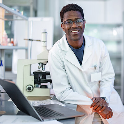 A scientist in a lab coat leans against a desk next to a microscope and poses for a photo.