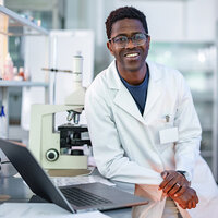 A scientist in a lab coat leans against a desk next to a microscope and poses for a photo.