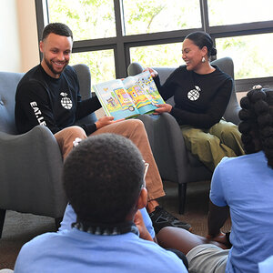 A seated couple in matching t-shirts read to a group of children—a photo of Stephen and Ayesha Curry. A seated couple in matching t-shirts read to a group of children—a photo of Stephen and Ayesha Curry.