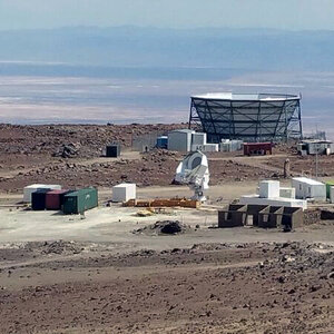 A large microwave telescope sits on a barren plain with mountains in the background.