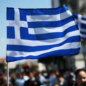 The blue and white national flag of Greece flies above a crowd at a parade. The blue and white national flag of Greece flies above a crowd at a parade.